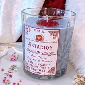 May include: A gray glass candle jar with a red and white label that reads "ATMALIS ASTARION Aged Brandy Bergamot & Rosemary Baldur's Gate 3 Inspired Candle Soy Wax HANDMADE".