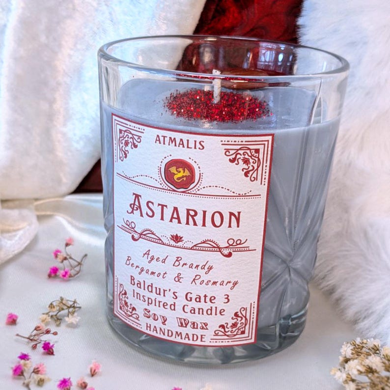 May include: A gray glass candle jar with a red and white label that reads "ATMALIS ASTARION Aged Brandy Bergamot & Rosemary Baldur's Gate 3 Inspired Candle Soy Wax HANDMADE".