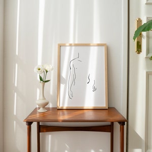Pu&ograve; includere: Un disegno a linea in bianco e nero incorniciato di un torso umano, con le braccia alzate, in stile minimalista. La cornice &egrave; in legno chiaro e il disegno &egrave; su carta bianca.