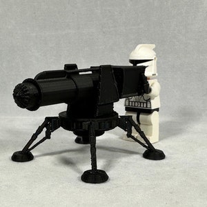 May include: A black 3D-printed artillery cannon with a detailed design, supported by a tripod base. A white and black toy soldier figure stands next to the cannon. The cannon has a textured barrel and intricate mechanical details.