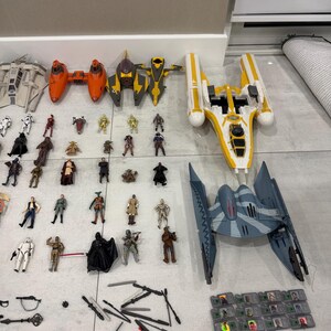以下が含まれることがあります： オレンジ、黄色、グレーの宇宙船モデルや、多数のキャラクターフィギュアを含む、スターウォーズのおもちゃのコレクション。おもちゃは明るい色の表面に配置されており、スターウォーズの世界観を代表する様々なキャラクターや乗り物を展示しています。