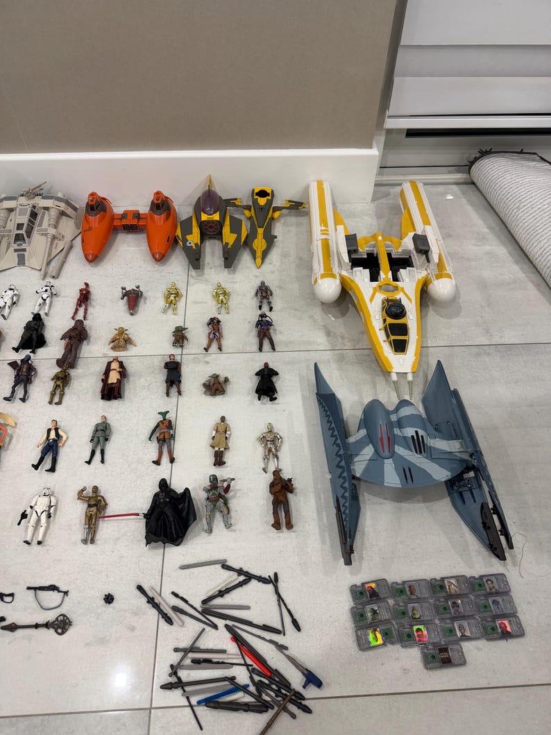 以下が含まれることがあります： オレンジ、黄色、グレーの宇宙船モデルや、多数のキャラクターフィギュアを含む、スターウォーズのおもちゃのコレクション。おもちゃは明るい色の表面に配置されており、スターウォーズの世界観を代表する様々なキャラクターや乗り物を展示しています。