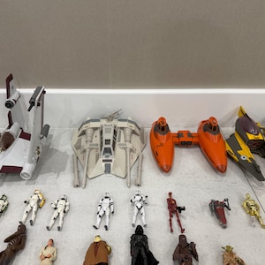 以下が含まれることがあります： スターウォーズのおもちゃのコレクション。白、オレンジ、黄色などの様々な宇宙船と、ストームトルーパー、ダース・ベイダー、ヨーダなどのキャラクターのアクションフィギュアがいくつかあります。おもちゃは白い表面に配置されています。