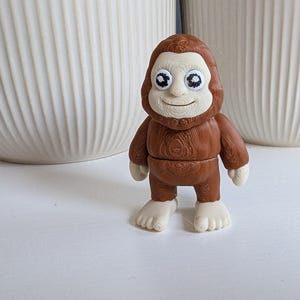May include: A small, brown Bigfoot figurine with a light-colored face, hands, and feet. The figurine has large, expressive eyes and a smiling expression. The body has a wood-grain texture. The figurine is standing on a white surface.