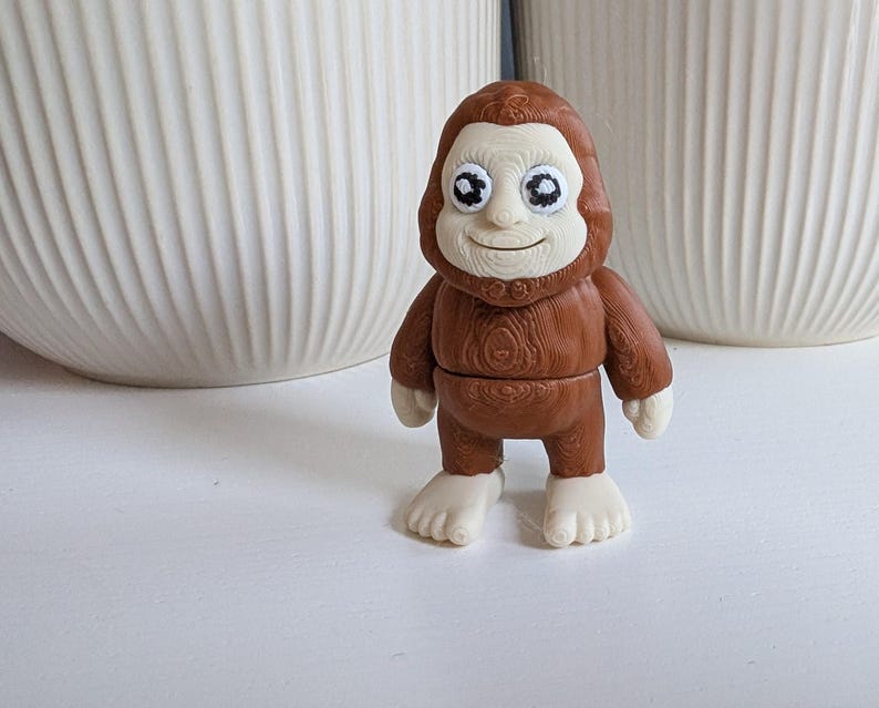 May include: A small, brown Bigfoot figurine with a light-colored face, hands, and feet. The figurine has large, expressive eyes and a smiling expression. The body has a wood-grain texture. The figurine is standing on a white surface.