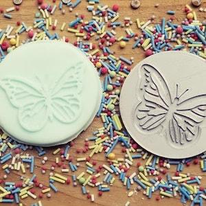 May include: Two round, decorative cookie cutters, one light blue and one gray, both featuring a butterfly design. The cutters are surrounded by colorful sprinkles.