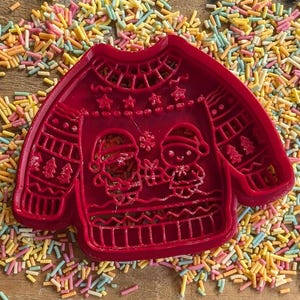 K&ouml;nnte beinhalten: Roter Kunststoff-Ausstecher in Form eines Weihnachtspullovers. Das Design des Pullevers zeigt Schneem&auml;nner, B&auml;ume und Schneeflocken. Der Ausstecher ist von bunten Streuseln umgeben, ideal zum Backen in der Weihnachtszeit.