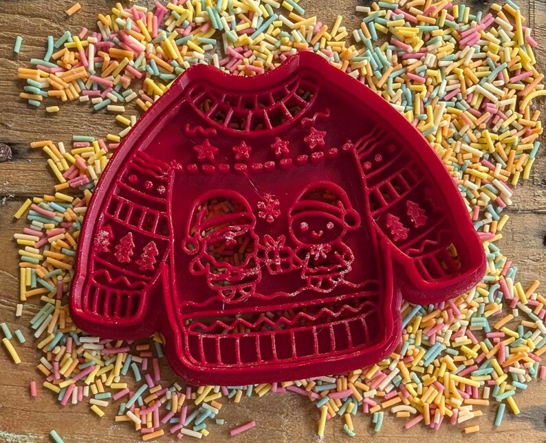 K&ouml;nnte beinhalten: Roter Kunststoff-Ausstecher in Form eines Weihnachtspullovers. Das Design des Pullevers zeigt Schneem&auml;nner, B&auml;ume und Schneeflocken. Der Ausstecher ist von bunten Streuseln umgeben, ideal zum Backen in der Weihnachtszeit.