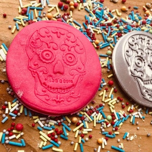Puede incluir: Dos decoraciones redondas de calaveras de az&uacute;car, una rosa brillante y otra plateada, est&aacute;n sobre una superficie de madera. Las calaveras de az&uacute;car est&aacute;n rodeadas de chispas de colores. La calavera rosa est&aacute; a la izquierda y la plateada a la derecha.