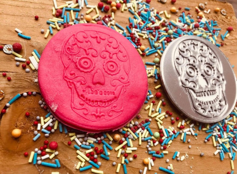 Puede incluir: Dos decoraciones redondas de calaveras de az&uacute;car, una rosa brillante y otra plateada, est&aacute;n sobre una superficie de madera. Las calaveras de az&uacute;car est&aacute;n rodeadas de chispas de colores. La calavera rosa est&aacute; a la izquierda y la plateada a la derecha.