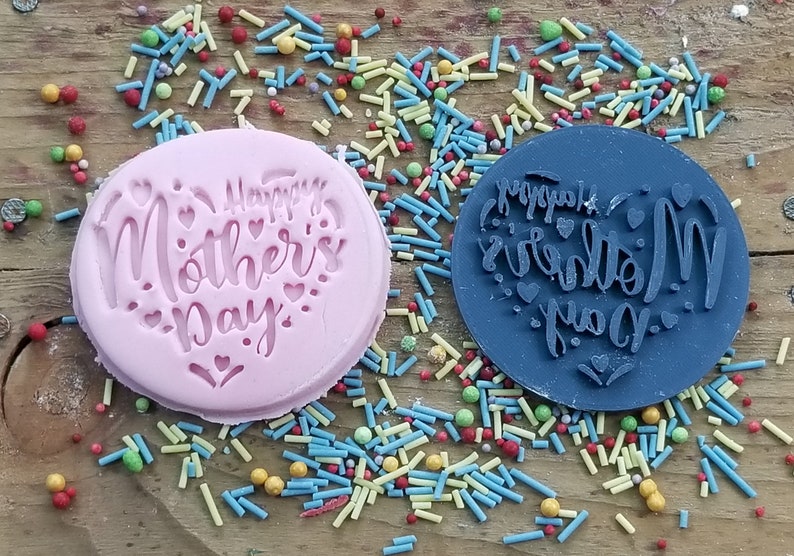 Puede incluir: Dos sellos de silicona, uno rosa y otro azul, con el texto "Happy Mother's Day" en forma de coraz&oacute;n. Los sellos est&aacute;n rodeados de espolvoreados coloridos.