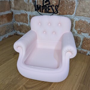 May include: A light pink miniature armchair-shaped tray with a tufted back and armrests. The tray is designed with a shallow basin for holding small items. The sign above the tray says "PRINT 2 IMPRESS".