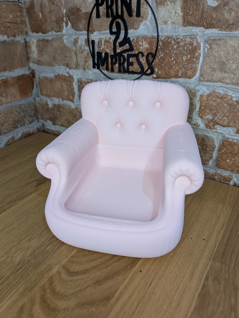 May include: A light pink miniature armchair-shaped tray with a tufted back and armrests. The tray is designed with a shallow basin for holding small items. The sign above the tray says "PRINT 2 IMPRESS".