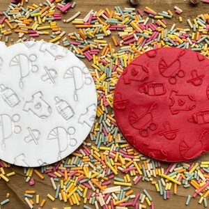 May include: Two round silicone stamps with baby-themed designs. One stamp is white with a light gray design and the other is red with a dark red design. The designs include a baby bottle, a pacifier, a baby carriage, and a onesie.