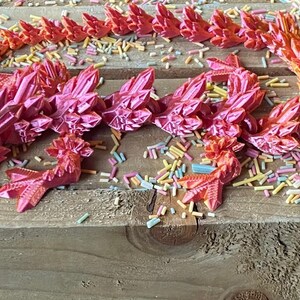 May include: A pink and orange 3D printed dragon figurine with a spiky, crystal-like design. The dragon is lying on a wooden surface with colorful sprinkles scattered around it.