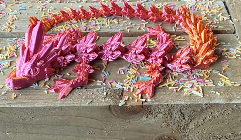 May include: A pink and orange 3D printed dragon figurine with a spiky, crystal-like design. The dragon is lying on a wooden surface with colorful sprinkles scattered around it.