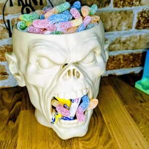 May include: A cream-colored, zombie-head-shaped candy bowl filled with colorful gummy candies. The zombie's mouth is open, revealing more candy. The bowl sits on a wooden surface, with a brick wall in the background.