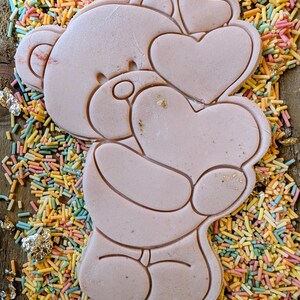 May include: A pale pink bear-shaped biscuit holding two heart-shaped biscuits. The biscuit is on a bed of colourful sprinkles. The bear has detailed outlines and is a charming treat.