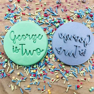 May include: Two green and gray round cookie cutters with the text "George is Two" and "George is Two" on them. The cookie cutters are surrounded by colorful sprinkles.