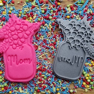 May include: Two cookie cutters in the shape of a mason jar with a floral bouquet on top. One is bright pink, the other is gray. The word "Mom" is written on the front of each. The cutters are surrounded by colorful sprinkles.