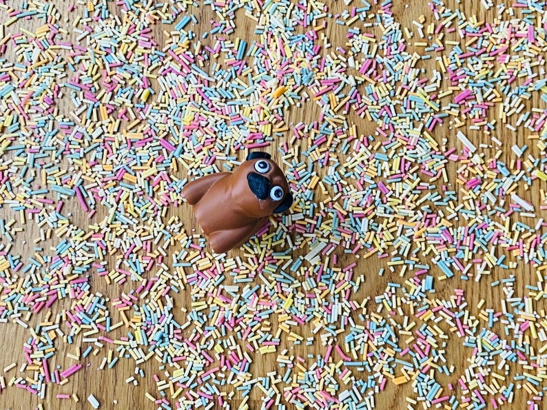 Op de afbeelding: Een bruin speelgoedpug met goggle-ogen is omringd door kleurrijke sprinkels op een houten oppervlak.