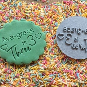 May include: A green cookie with the text "Ava-grace is 3 Three" and a heart, next to a gray stamp with the same text in reverse. The items are surrounded by colorful sprinkles, perfect for a birthday celebration.