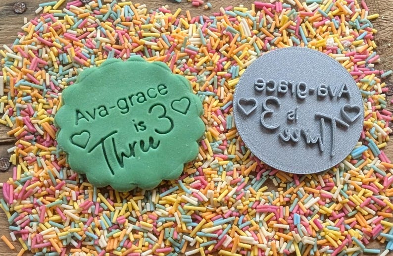 May include: A green cookie with the text "Ava-grace is 3 Three" and a heart, next to a gray stamp with the same text in reverse. The items are surrounded by colorful sprinkles, perfect for a birthday celebration.