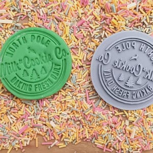 Może przedstawiać: Dwa okrągłe stemple do ciasteczek, zielony i szary, z tekstem "North Pole Milk & Cookie Co. Baking Fresh Daily".