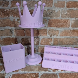 May include: A set of three light purple organizers. The set includes a crown-shaped stand, a two-compartment holder, and a tiered lipstick holder. The word "PLOUISE" is embossed on each item.