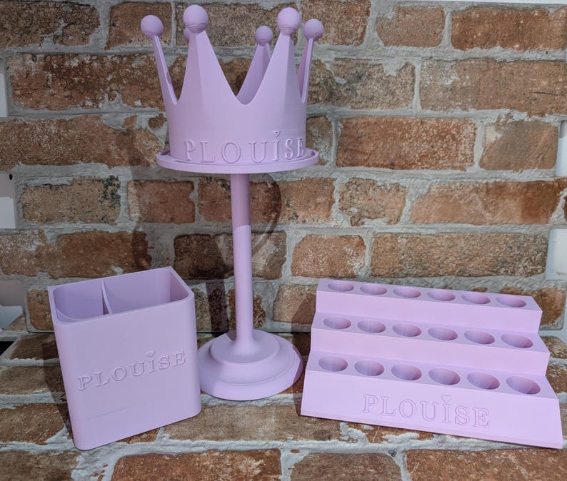 May include: A set of three light purple organizers. The set includes a crown-shaped stand, a two-compartment holder, and a tiered lipstick holder. The word "PLOUISE" is embossed on each item.