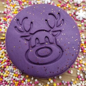 May include: Purple silicone stamp featuring a reindeer design. The reindeer has antlers, eyes, and a nose. The stamp is covered in glitter.