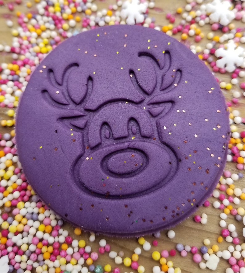 May include: Purple silicone stamp featuring a reindeer design. The reindeer has antlers, eyes, and a nose. The stamp is covered in glitter.