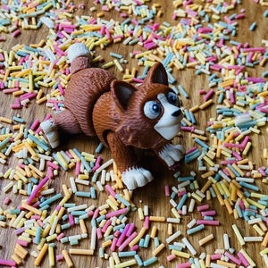 May include: A brown and white toy cat with large eyes and a playful expression is surrounded by colorful sprinkles on a wooden surface.