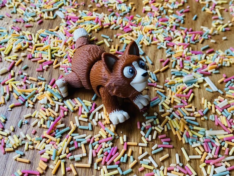 May include: A brown and white toy cat with large eyes and a playful expression is surrounded by colorful sprinkles on a wooden surface.