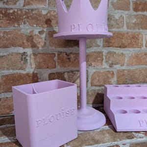 May include: A light purple, three-piece set of decorative organizers. The set includes a crown-shaped stand, a rectangular container, and a tiered tray. The word "PLouise" is embossed on the stand and container.