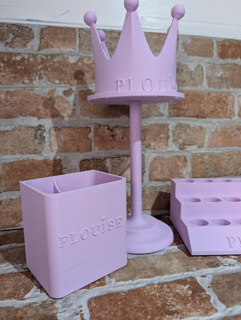 May include: A light purple, three-piece set of decorative organizers. The set includes a crown-shaped stand, a rectangular container, and a tiered tray. The word "PLouise" is embossed on the stand and container.