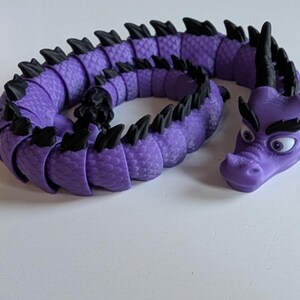 K&ouml;nnte beinhalten: Ein lila und schwarzes, segmentiertes Drachenspielzeug mit einer strukturierten Oberfl&auml;che. Der Drache hat einen gro&szlig;en Kopf mit schwarzen Augen und einer schwarzen Augenbraue.