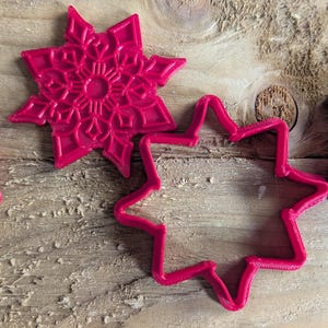 May include: Four bright pink cookie cutters in snowflake and star shapes are arranged on a wooden surface. The cutters have detailed designs and are made of plastic. The image is a close-up shot.