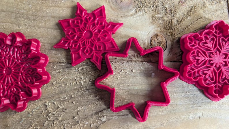 May include: Four bright pink cookie cutters in snowflake and star shapes are arranged on a wooden surface. The cutters have detailed designs and are made of plastic. The image is a close-up shot.