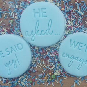 May include: Three light blue round cookies with white icing and blue lettering. The cookies say "He asked...", "She said yes!" and "We're engaged".