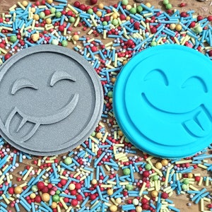 May include: Two round cookie cutters with a smiling face design. One is gray, the other is bright blue. The cutters are surrounded by colorful sprinkles, suggesting a baking or crafting theme. The design features a tongue sticking out.