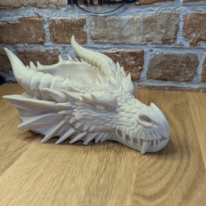 May include: A detailed, off-white dragon head sculpture with horns, sharp teeth, and textured scales. The dragon head is resting on a wooden surface. The word "IMPRESS" is visible in the background.