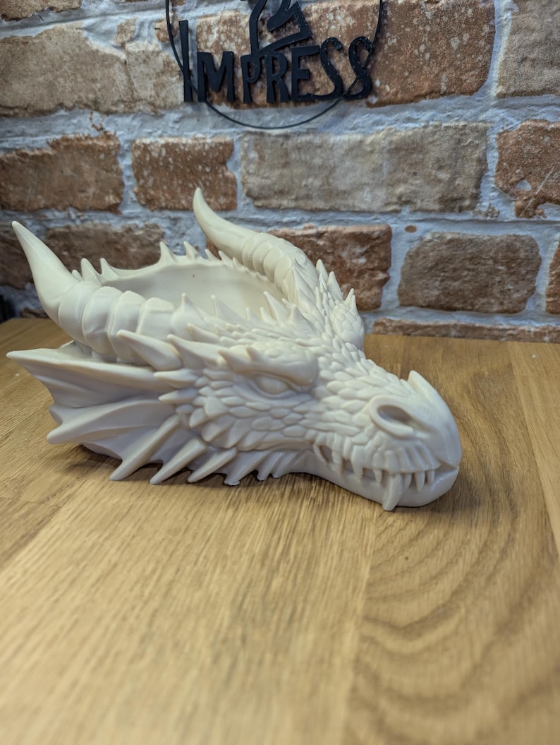 May include: A detailed, off-white dragon head sculpture with horns, sharp teeth, and textured scales. The dragon head is resting on a wooden surface. The word "IMPRESS" is visible in the background.