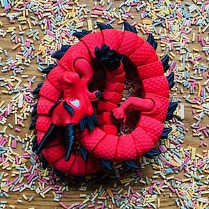 May include: A red plastic dragon toy curled up on a wooden surface covered in colorful sprinkles.