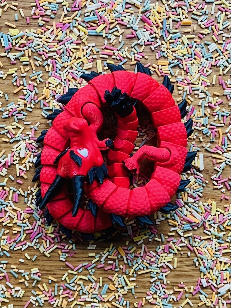 May include: A red plastic dragon toy curled up on a wooden surface covered in colorful sprinkles.