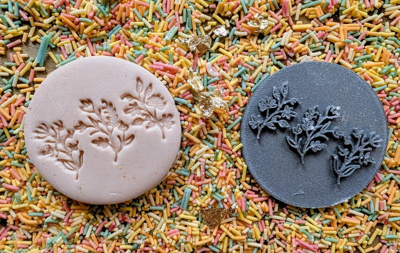 May include: Two round floral design stamps, one light pink and one dark gray, rest on a bed of colorful sprinkles. The stamps feature detailed botanical patterns, ideal for crafting or baking projects.