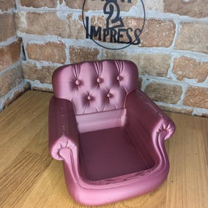 May include: A small, mauve-colored miniature armchair with a tufted back and rolled arms. The chair is a decorative item, possibly a trinket dish or display piece. The background includes a brick wall and the text "PRINT 2 IMPRESS".