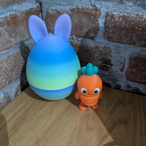 K&ouml;nnte beinhalten: Eine farbenfrohe, gradientenfarbene, eif&ouml;rmige Hasenfigur mit aufrechten Ohren, neben einer orangefarbenen Karottenfigur mit gr&uuml;nem Oberteil. Die Figuren stehen auf einer Holzoberfl&auml;che, mit einer Backsteinmauer im Hintergrund.