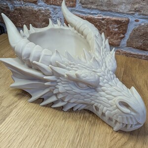 May include: A cream-colored dragon head planter with detailed scales, horns, and a wide open mouth. The planter has a shallow bowl-shaped opening on top for plants or other decorative items. The dragon head is resting on a wooden surface.