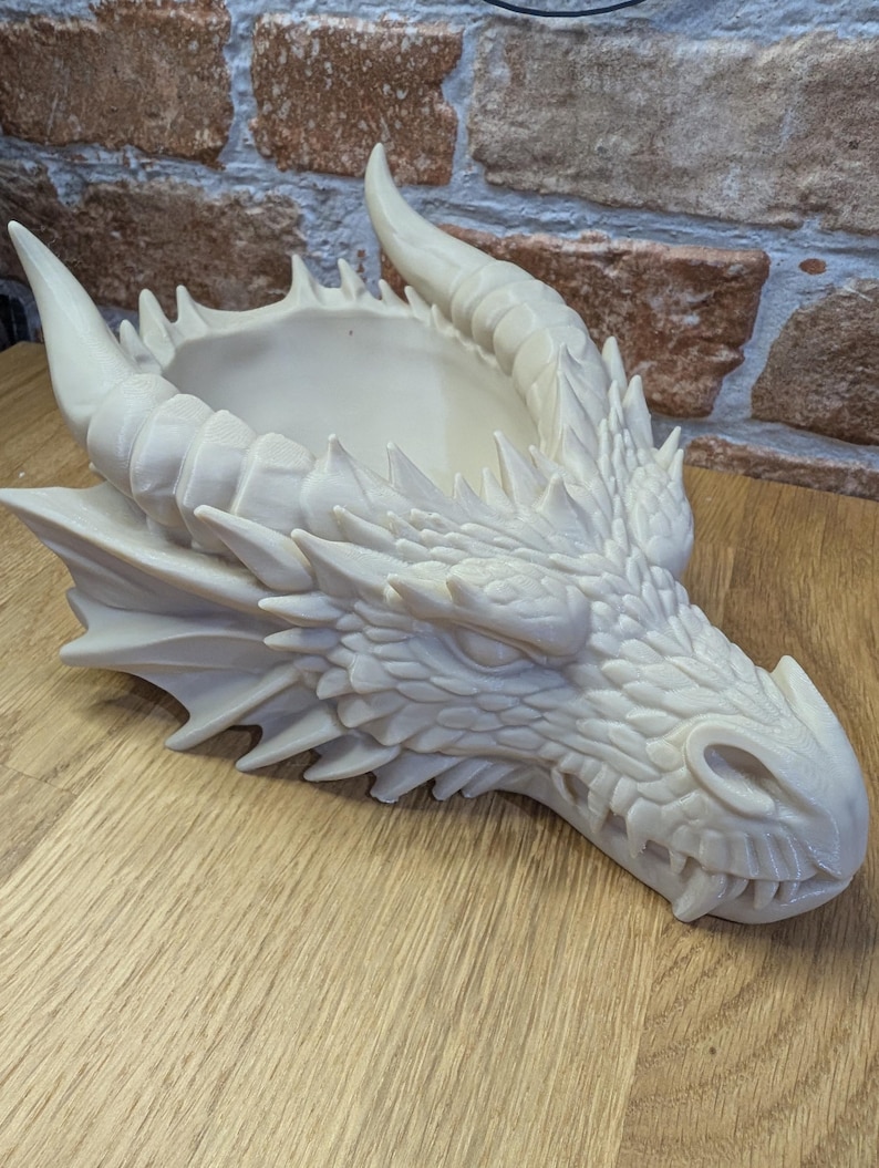 May include: A cream-colored dragon head planter with detailed scales, horns, and a wide open mouth. The planter has a shallow bowl-shaped opening on top for plants or other decorative items. The dragon head is resting on a wooden surface.